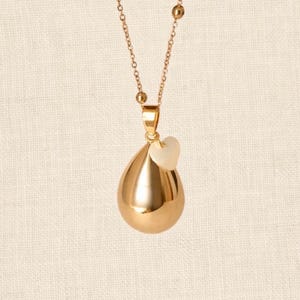 Peut inclure: Un collier pendentif doré avec un charm en forme de goutte et un petit cœur. La chaîne est ornée de petites perles rondes. Le pendentif est très réfléchissant, sur fond beige neutre.