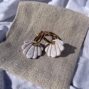 May include: A pair of gold hoop earrings with white seashell charms. The seashell charms have a speckled, tan pattern. The earrings are displayed on a beige linen pouch, set against a white background.