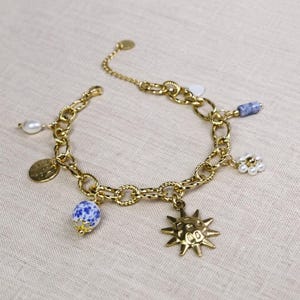 Può includere: Bracciale con ciondoli dorato con un ciondolo a forma di sole, una perla maculata blu e bianca, una perla e altri ciondoli decorativi. Il bracciale è su una superficie neutra.