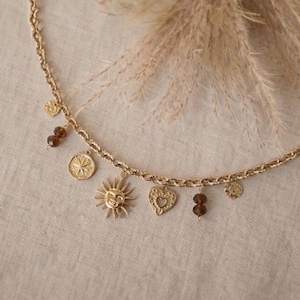 Op de afbeelding: Goudkleurige ketting met diverse bedels, waaronder zon-, hart- en muntvormige hangers. De ketting heeft ook kleine bruine kralen. De ketting heeft een textuurontwerp. De achtergrond is een neutraal beige.