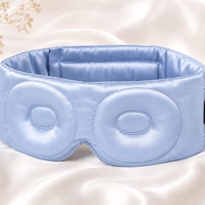 May include: A light blue satin sleep mask with two padded eye covers. The mask has a wide band and a small tag on the side. The background is a white satin fabric. The mask is designed to block out light for better sleep.