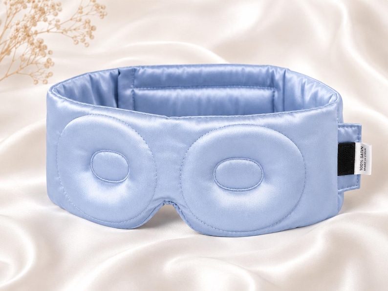 May include: A light blue satin sleep mask with two padded eye covers. The mask has a wide band and a small tag on the side. The background is a white satin fabric. The mask is designed to block out light for better sleep.