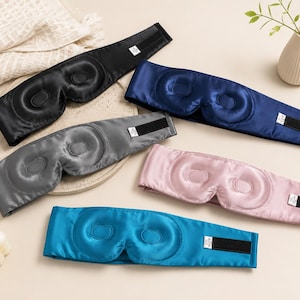 May include: A collection of satin sleep masks in various colors: black, navy blue, gray, pink, and teal. Each mask features a contoured design for the eyes and an adjustable Velcro strap for a secure fit. The masks are designed to block out light.