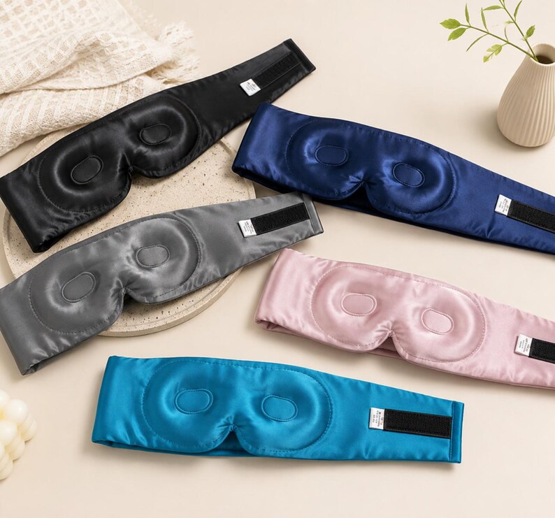 May include: A collection of satin sleep masks in various colors: black, navy blue, gray, pink, and teal. Each mask features a contoured design for the eyes and an adjustable Velcro strap for a secure fit. The masks are designed to block out light.