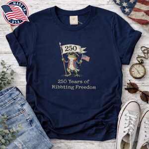 Könnte beinhalten: Marineblaues T-Shirt mit einer Cartoon-Frosch-Illustration in Kolonialkleidung, der eine Flagge mit der Zahl 250 und eine amerikanische Flagge hält. Der Text "250 Years of Ribbiting Freedom" ist darunter gedruckt. Das T-Shirt hat einen Rundhalsausschnitt.