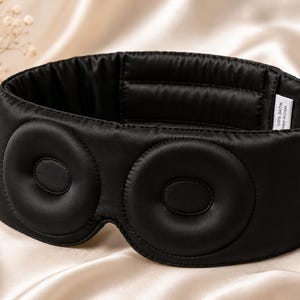 May include: A black satin sleep mask with two circular eye covers. The mask is designed to block out light and promote relaxation. The mask is made of 100% satin.