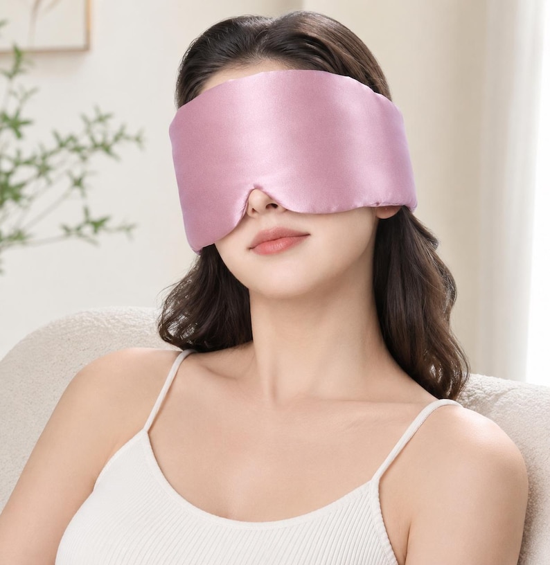 May include: A light pink silk eye mask covers the eyes of a person. The mask is made of a smooth, shiny fabric and has a curved shape to fit the face. The person is wearing a white tank top. The background is blurred.