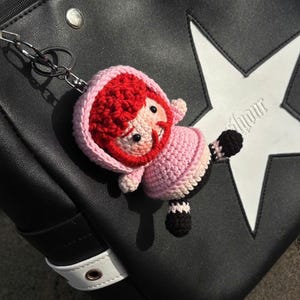 May include: A handmade crochet doll keychain with red hair, a pink outfit, and black and white striped legs. The doll is attached to a black handbag with a white star design.