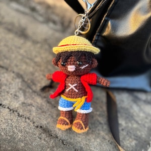 May include: Crocheted keychain featuring a character with dark skin, wearing a yellow straw hat, red jacket, blue shorts, and brown boots. The character has a scar under the eye and a cross-shaped chest detail. The keychain is attached to a black bag.