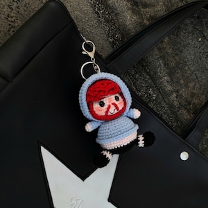 Könnte beinhalten: Ein handgefertigter Häkel-Schlüsselanhänger mit einer kleinen Figur mit rotem Bart und blauem Kapuzenpullover. Der Schlüsselanhänger ist an einer schwarzen Tragetasche mit weißem Stern-Design und dem geprägten Wort "Flyour" befestigt.