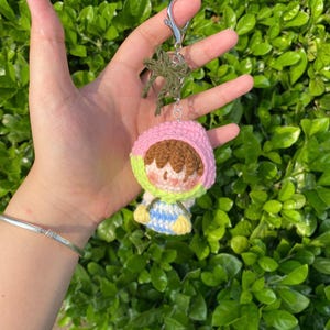 Op de afbeelding: Een handgemaakte gehaakte poppen sleutelhanger met een klein figuur met een roze capuchon, bruin haar en een blauw en gele outfit. De sleutelhanger heeft een zilveren sluiting en een groen gehaakt accent. De pop wordt tegen een achtergrond van groen gebladerte gehouden.