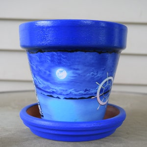 Op de afbeelding: Blauw geverfde terracotta bloempot met een nautische scène van een maan, wolken en een scheepsrad. De pot heeft een bijpassende blauwe schotel.