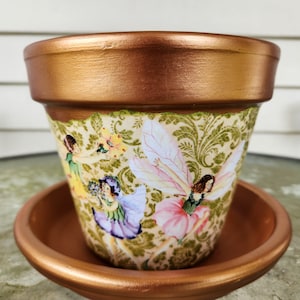 Peut inclure: Pot de fleur en terre cuite avec un bord et une soucoupe de couleur cuivre. Le pot est décoré d'un papier à motifs représentant des fées aux robes et aux ailes colorées. Le motif comprend des éléments floraux et des volutes.