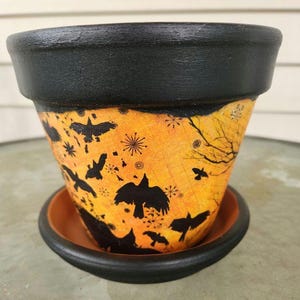 May include: A decorative terracotta flower pot with a black rim and saucer. The pot features a vibrant orange background with black silhouettes of crows, branches, and snowflakes. The pot is designed for planting flowers or herbs.