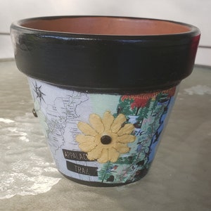 May include: A terracotta flower pot painted black with a map of the Appalachian Trail, a yellow flower, and the words "Appalachian Trail" on the side.