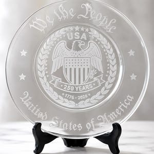 Puede incluir: Plato de vidrio transparente con diseño grabado. Presenta las palabras "We the People" y "United States of America". Un emblema de águila y el texto "250 YEARS 1776-2026" también están grabados. El plato se exhibe sobre un soporte negro.