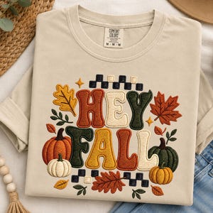 Könnte beinhalten: Ein beiges Kurzarm-T-Shirt mit einem gestickten Design, das den Text HEY FALL in großen, farbenfrohen Buchstaben zeigt. Die Grafik enthält orange, grüne und weiße Kürbisse, Herbstblätter und ein schwarz-weißes Karomuster als Umrandung.