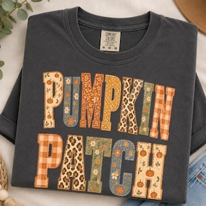 Puede incluir: Una camiseta de cuello redondo gris carbón con el texto PUMPKIN PATCH en letras grandes. Cada letra está rellena con un patrón diferente, incluyendo calabazas, estampado de leopardo, cuadros vichy naranjas y diseños florales sobre tela oscura.