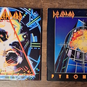 Könnte beinhalten: Zwei Def Leppard Albumcover. Das Album "Hysteria" zeigt ein Gesicht in einem Dreieck, mit dem Albumtitel darunter. Das Albumcover "Pyromania" zeigt ein Ziel über einem Gebäude mit dem Albumtitel darunter. Beide Cover haben das Def Leppard-Logo.