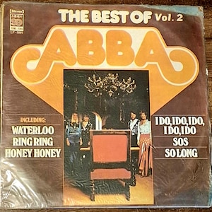 Könnte beinhalten: Ein quadratisches Vinyl-Schallplattencover mit dem Titel The Best of ABBA Vol. 2. Das Cover zeigt ein großes orangefarbenes ABBA-Logo, ein zentrales Foto der Bandmitglieder an einem Stuhl und eine Liste mit Liedern wie Waterloo, Ring Ring und SOS.