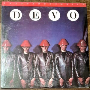 May include: Album cover featuring the band Devo. The band members are wearing silver jackets, black shirts, and red, tiered hats. The album title, "Freedom of Choice," is at the top. The band's name, "DEVO," is in large white circles.