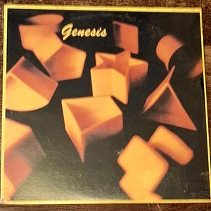 Op de afbeelding: Albumhoes met een zwarte achtergrond en verspreide oranje geometrische vormen. Het woord "Genesis's" staat in gouden letters bovenaan. De albumhoes heeft een gele rand.