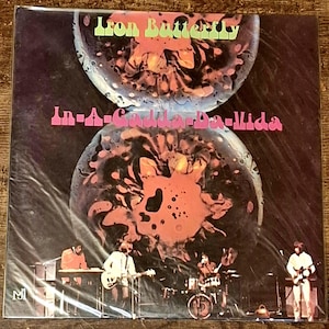 May include: A vinyl record album cover for Iron Butterfly's In-A-Gadda-Da-Vida. The cover features two large, circular, abstract orange and black shapes against a dark background. Below, a band performs with instruments on a stage.