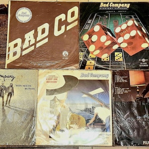 May include: A collection of vintage vinyl record albums by the band Bad Company. The album covers feature various designs, including the band's logo, dice, and band members. The albums are displayed on a wooden surface.