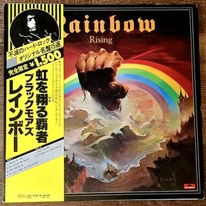 May include: Rainbow "Rising" vinyl record album cover. The album art features a fist emerging from a sea of clouds, with a rainbow arching above. The album title is in gold lettering. A yellow sticker with Japanese text is on the left.