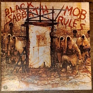 May include: Album cover for Black Sabbath's "Mob Rules." The artwork features a dark, gritty scene with figures in hooded cloaks, a tattered cloth, and the band's name in a distressed font. The overall color palette is muted with browns and grays.