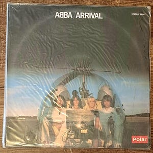 Könnte beinhalten: Ein Schallplattencover für ABBA Arrival mit einem Foto von vier Personen, die im Cockpit eines Hubschraubers sitzen. Der Text ABBA ARRIVAL ist oben in Weiß gedruckt, mit STEREO AB267 in der Ecke und einem roten Polar-Logo am unteren Rand.