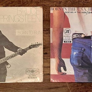 Può includere: Due copertine di dischi in vinile vintage. La prima copertina è in bianco e nero, con Bruce Springsteen con una chitarra, e il testo "BRUCE SPRINGSTEEN" e "BORN TO RUN". La seconda copertina ha uno sfondo a strisce rosse e bianche, con il testo "BORN IN THE U.S.A." e "DANCING IN THE DARK".