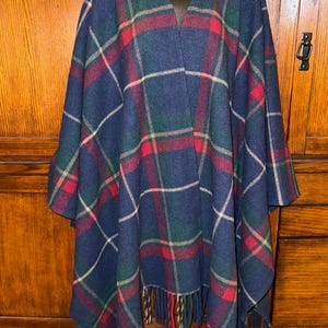 May include: A navy blue poncho featuring a plaid pattern in shades of forest green, bright red, and cream. The garment has an open front design, a V-neckline, and a hemline finished with long, thick fringe tassels along the bottom edge.