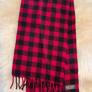 May include: A red and black checked scarf with a fringed edge. The scarf displays a traditional buffalo check pattern. The label reads "THE GAP".