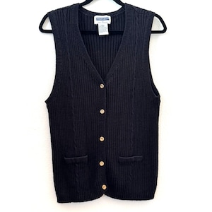 以下が含まれることがあります： Vネックと金色のボタンが付いた、黒色のノースリーブセーターベスト。 ベストには2つのフロントポケットとリブ編みのデザインがあります。 衣類は白い背景に対して黒いハンガーに吊るされています。