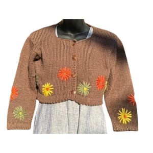May include: Brown, cropped cardigan sweater with embroidered floral details in orange, yellow, and green. The sweater has a round neckline, long sleeves, and button closures. The floral embroidery is on the front and sleeves.