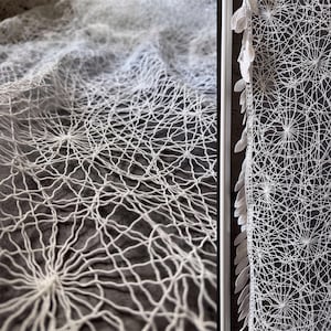 May include: A white lace fabric featuring an intricate, web-like geometric pattern with radiating circular centres. The edge of the textile is finished with a decorative border of small, leaf-shaped white fabric appliques attached in a vertical row.