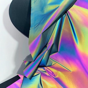 May include: Abstract composition featuring a dark, semi-circular shape juxtaposed with iridescent, folded fabric. The fabric displays a spectrum of colors, including blues, greens, yellows, and purples, creating a dynamic visual effect. The artwork is set against a white background.