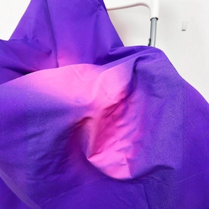 Puede incluir: Primer plano de una tela con un degradado de color. La tela pasa de un morado intenso en los bordes a un rosa vibrante en el centro. La tela está drapeada sobre una silla blanca.