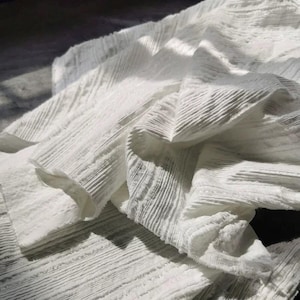 May include: Close-up of white fabric with a textured, ribbed design. The fabric is layered and folded, creating shadows and highlights. The material appears soft and lightweight, suitable for clothing or home decor.