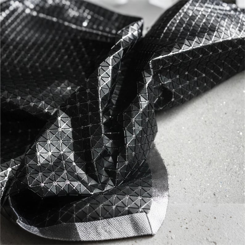 May include: A black fabric featuring a reflective, geometric, three-dimensional triangular pattern. The material has a glossy, metallic finish that catches the light, creating a textured, faceted appearance across the folded surface of the textile.