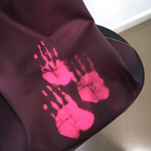 May include: A dark burgundy fabric with three bright pink handprints. The handprints are arranged in a vertical pattern, with the largest at the bottom and the smallest at the top. The fabric appears to be draped over an unseen object.