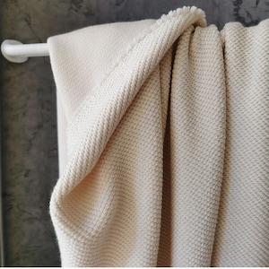 Pode incluir: Uma manta creme texturizada drapeada sobre uma barra de toalha branca. A manta tem uma trama tipo waffle e uma borda com franjas. A cor neutra e a textura sugerem um item de decoração acolhedor.