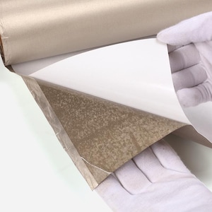 May include: A roll of beige paper being unrolled to reveal a white surface. The paper is held by gloved hands, showing the texture and color contrast between the two sides. The paper appears to be new and unused.