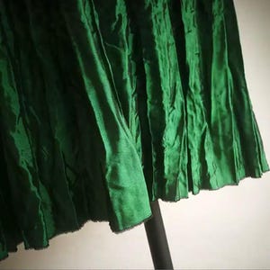 Pode incluir: Um close-up de uma saia plissada verde esmeralda. O tecido tem uma aparência texturizada com dobras verticais e uma bainha ligeiramente desfiada. A saia está pendurada em um suporte preto, com fundo branco.