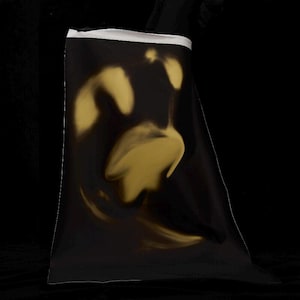 Puede incluir: Un bolso rectangular de tela negra que presenta un diseño abstracto en tono dorado realizado con aerógrafo en la parte frontal. El bolso tiene una banda horizontal blanca distintiva a lo largo del borde superior, creando un contraste.