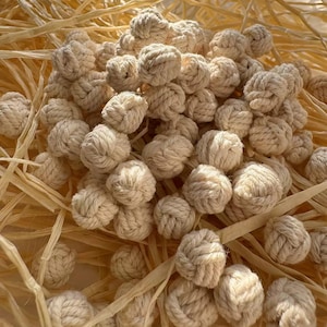 May include: A collection of off-white, knotted rope balls. The balls are tightly wound and clustered together, resting on a bed of light brown, dried plant fibers. The lighting creates soft shadows, highlighting the texture of the rope and fibers.