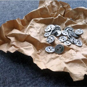 Op de afbeelding: Een verzameling kleine, zilverkleurige metalen knopen met elk twee gaatjes, verspreid op een gekreukeld vel bruin papier. Het papier rust op een donkergrijze ondergrond, wat een contrast in texturen en kleuren creëert. De knopen lijken vintage of antiek.