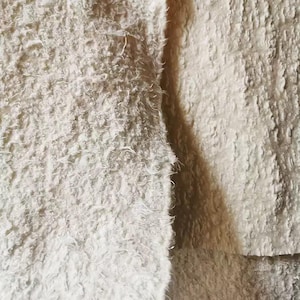 Peut inclure: Gros plan sur deux sections de tissu blanc cassé texturé. La section de gauche a un aspect hirsute et duveteux, tandis que la section de droite est plus lisse avec une texture légèrement côtelée. L'éclairage crée des ombres et des reflets.
