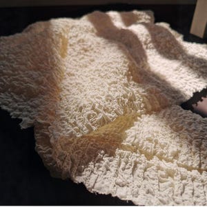 Könnte beinhalten: Nahaufnahme eines strukturierten, cremefarbenen Stoffes mit zerknittertem Aussehen. Der Stoff wird von Sonnenlicht beleuchtet, wodurch Schatten entstehen und die detaillierte Oberfläche hervorgehoben wird. Das Material scheint weich zu sein und kann für Kleidung oder Heimdekoration verwendet werden.